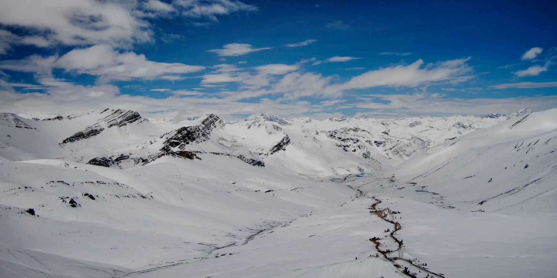 A winding trekking path through a vast snow-covered mountain valley under a bright blue sky.