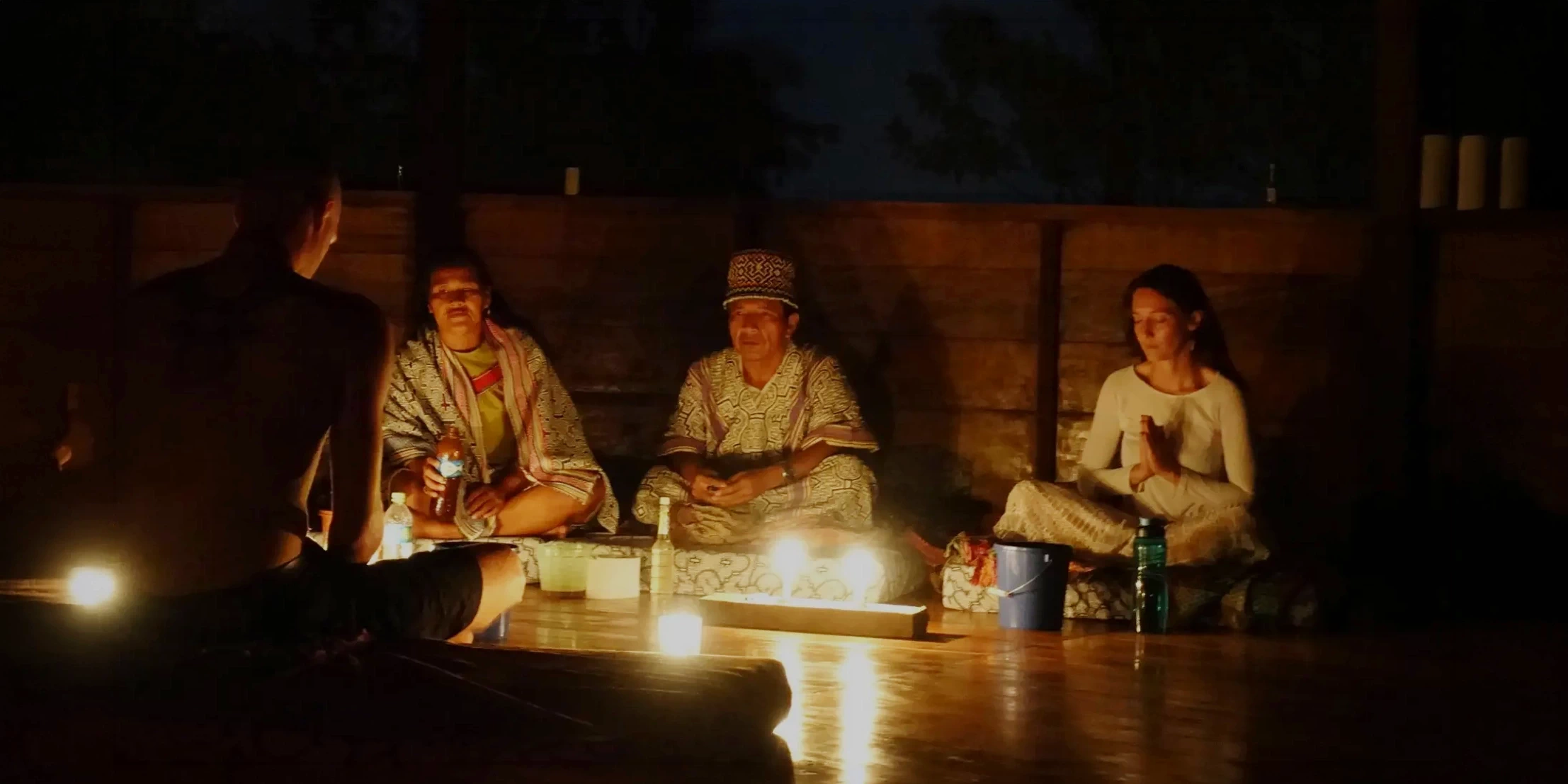 An Ayahuasca night ceremony in the Amazon rainforest with a shaman and participants sitting around candles in a traditional wooden maloka.