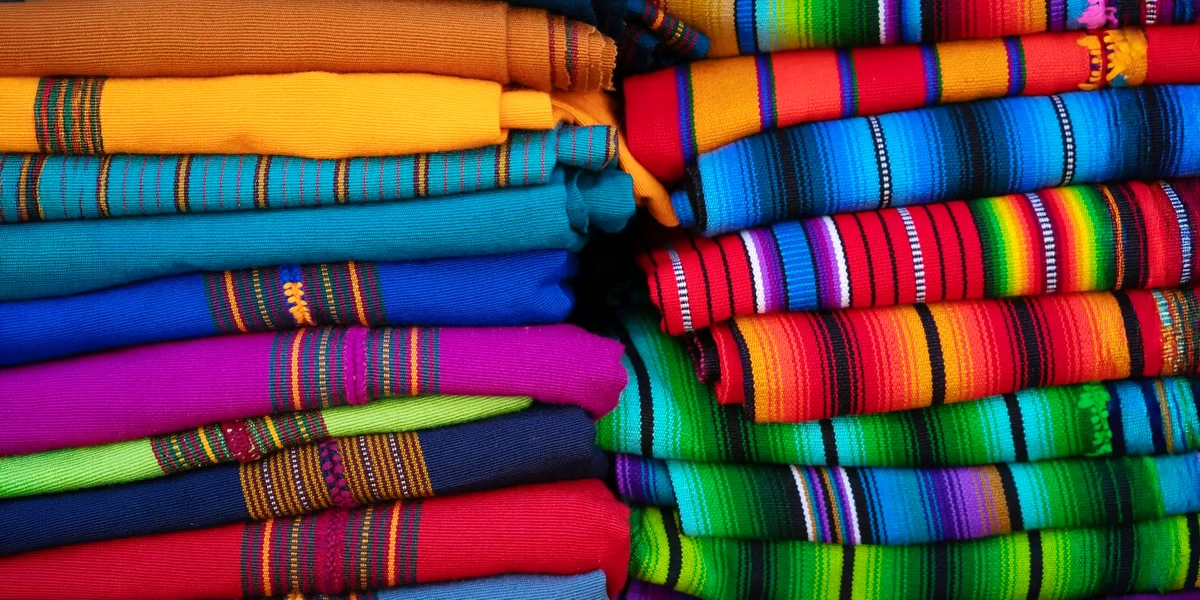 A close-up stack of vibrant, folded Peruvian textiles featuring traditional Andean geometric patterns and bright colors.