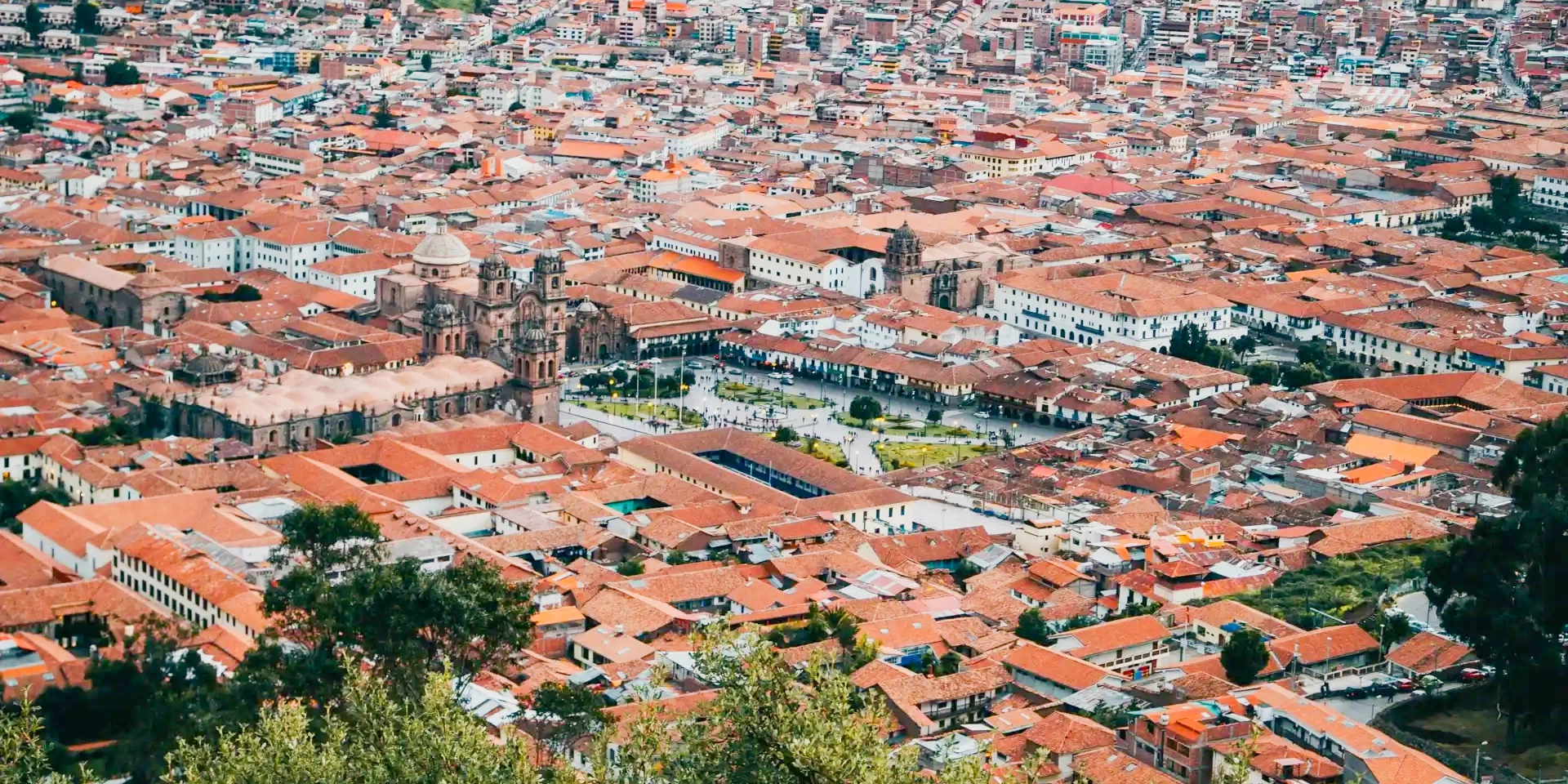 Wide panoramic view of Cusco city showing the iconic red-tiled roofs and historic churches under a bright sky.