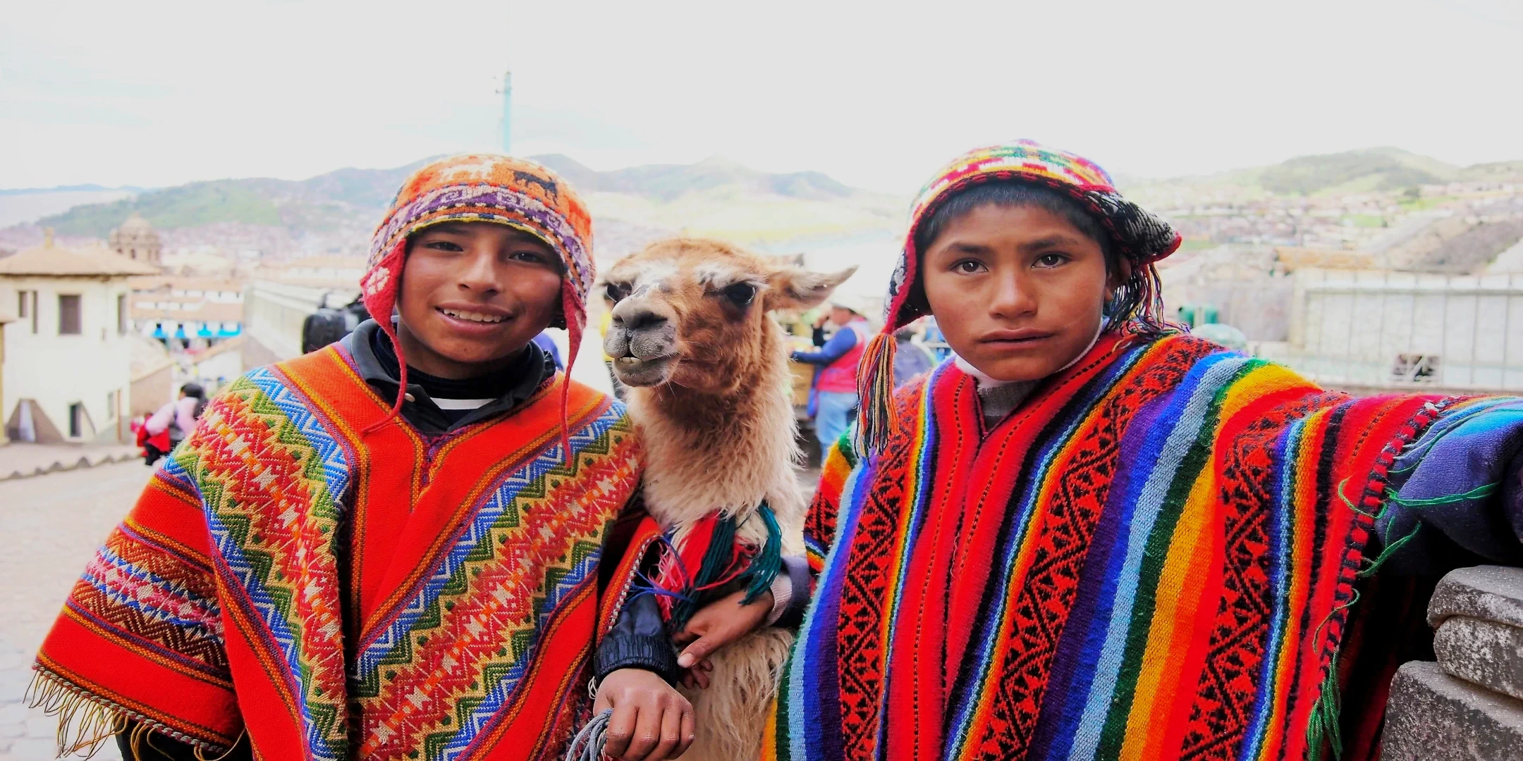 Two smiling local children wearing vibrant traditional ponchos and chullos, posing with a friendly alpaca.