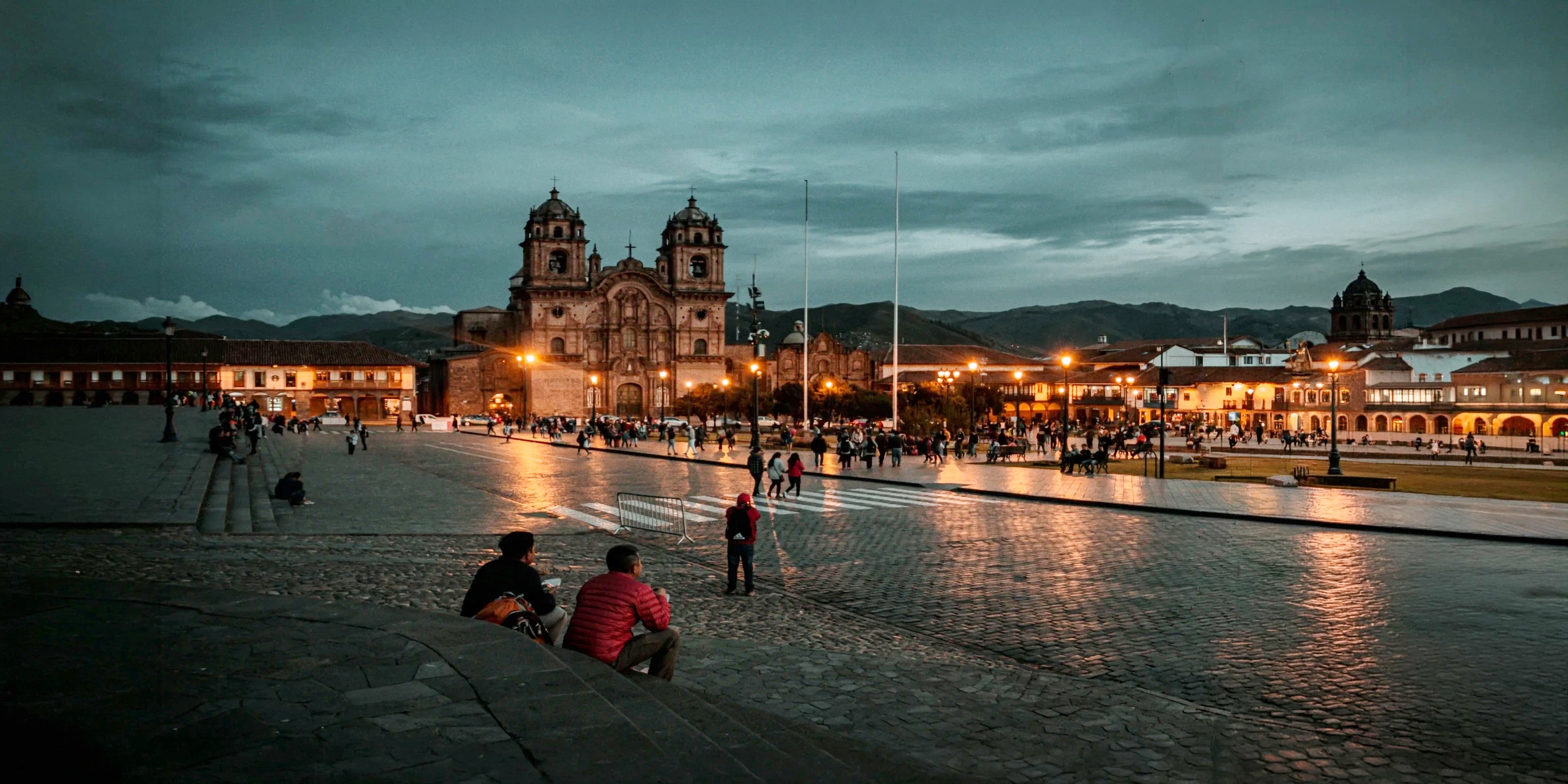 The illuminated Plaza de Armas in Cusco at night, featuring the Cathedral and people sitting on the steps under a dark sky.