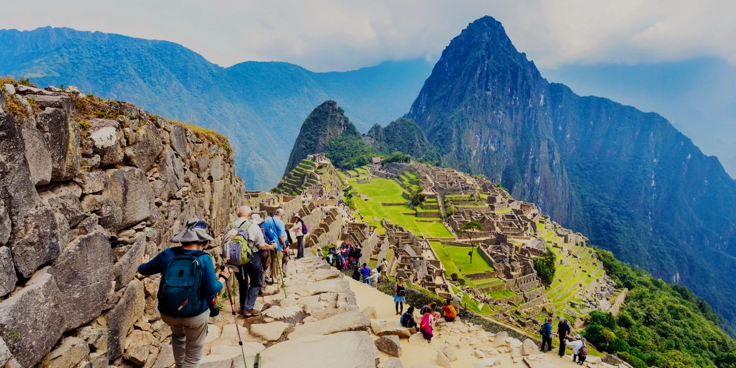 A group of tourists with backpacks and trekking poles walking down a stone path towards the ancient ruins of Machu Picchu.