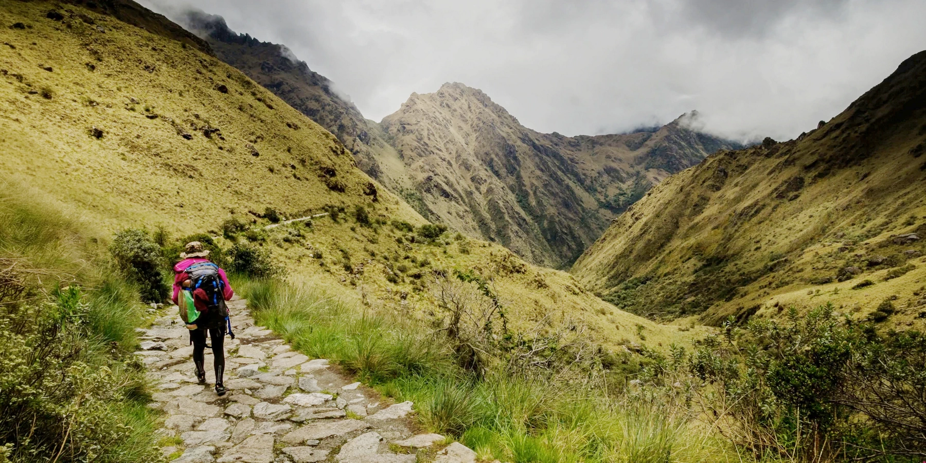 A person with a large backpack walking on an ancient stone Inca path surrounded by vast mountain valleys.
