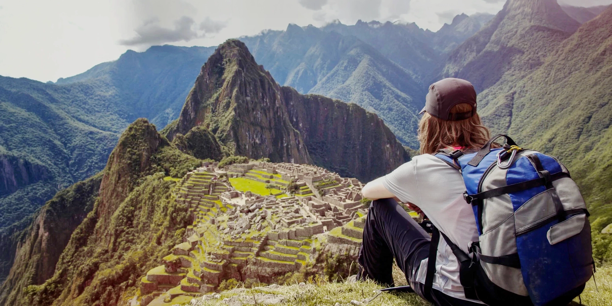 A hiker with a backpack sitting on a hill and looking out over the majestic Machu Picchu citadel and surrounding peaks. 