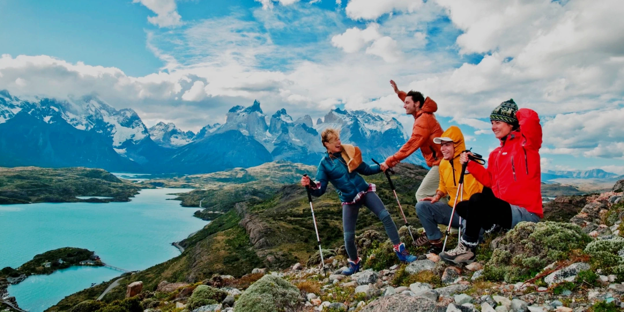 A group of four cheerful hikers sitting on a rocky ridge overlooking a turquoise glacial lake and snowy mountain peaks.