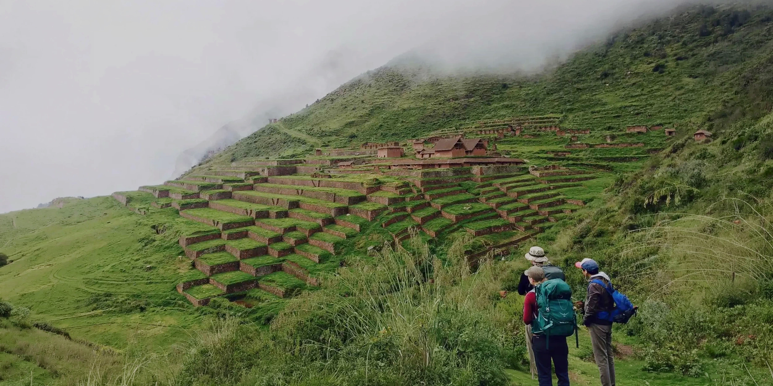 Hikers standing on a mountain path overlooking the extensive stone agricultural terraces and ruins of Huchuy Qosqo under a misty sky.