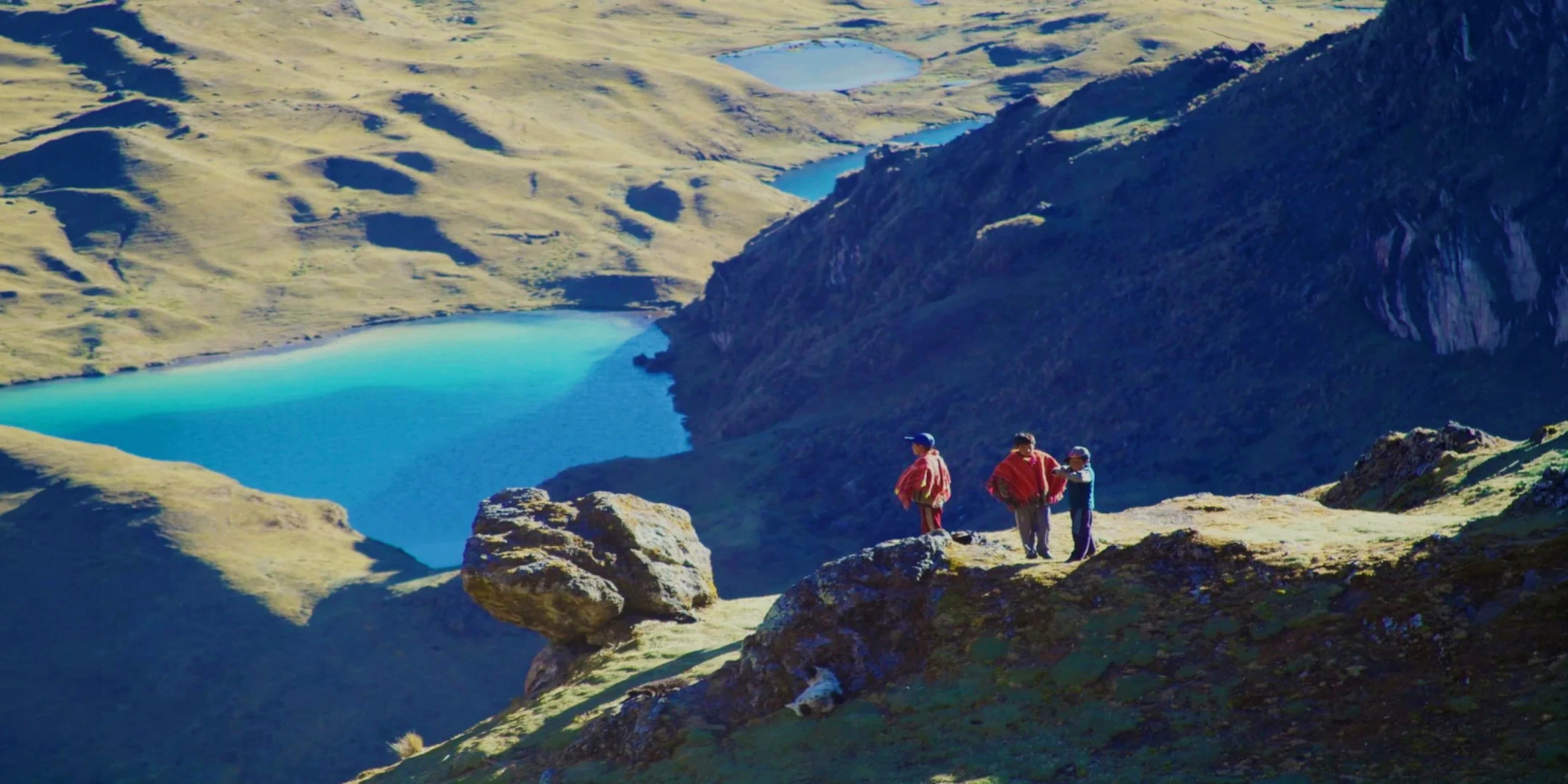 Local Andean children in traditional ponchos standing on a rocky outcrop overlooking a large turquoise lagoon.