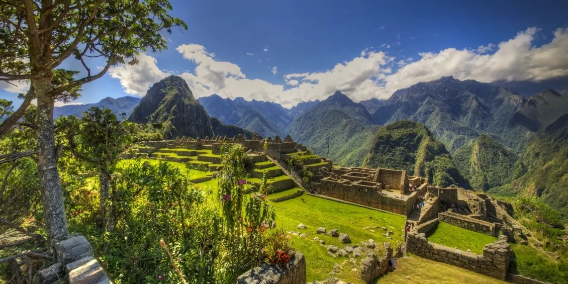 A bright, sunny panoramic view of the Machu Picchu citadel with green terraces and the Huayna Picchu mountain in the background.