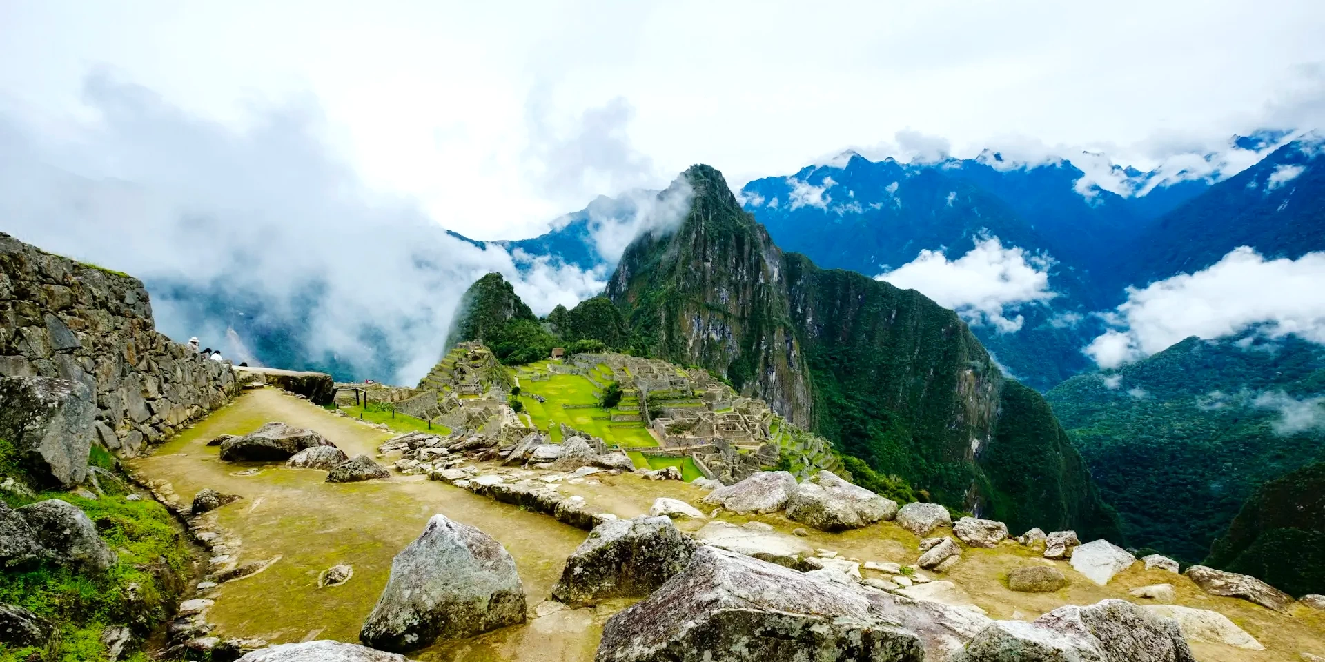 A wide-angle landscape view of Machu Picchu showing the stone trails, the main citadel, and the surrounding canyon mountains under a cloudy sky.
