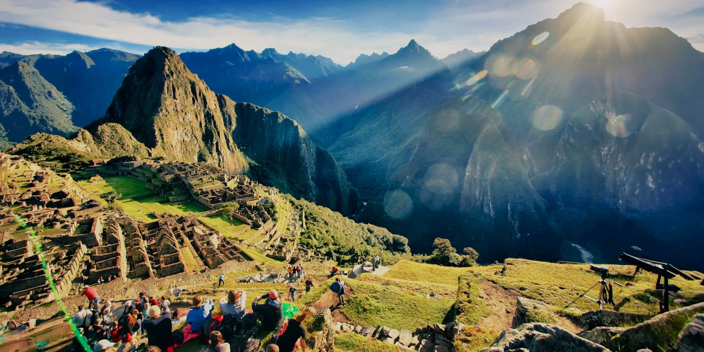 Panoramic sunrise view of the Machu Picchu citadel with tourists watching from the terraces and Huayna Picchu mountain in the background.