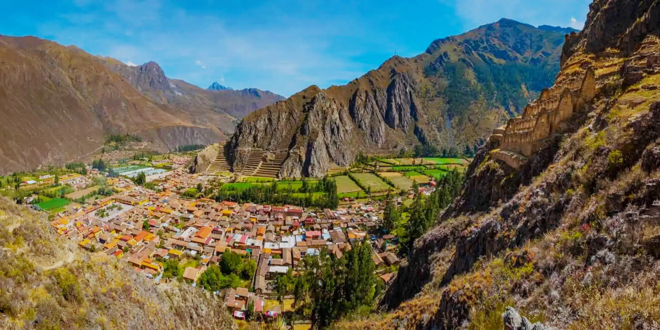Panoramic view of the historic town of Ollantaytambo in the Sacred Valley, showing the traditional red-roofed houses nestled at the foot of massive Inca ruins.