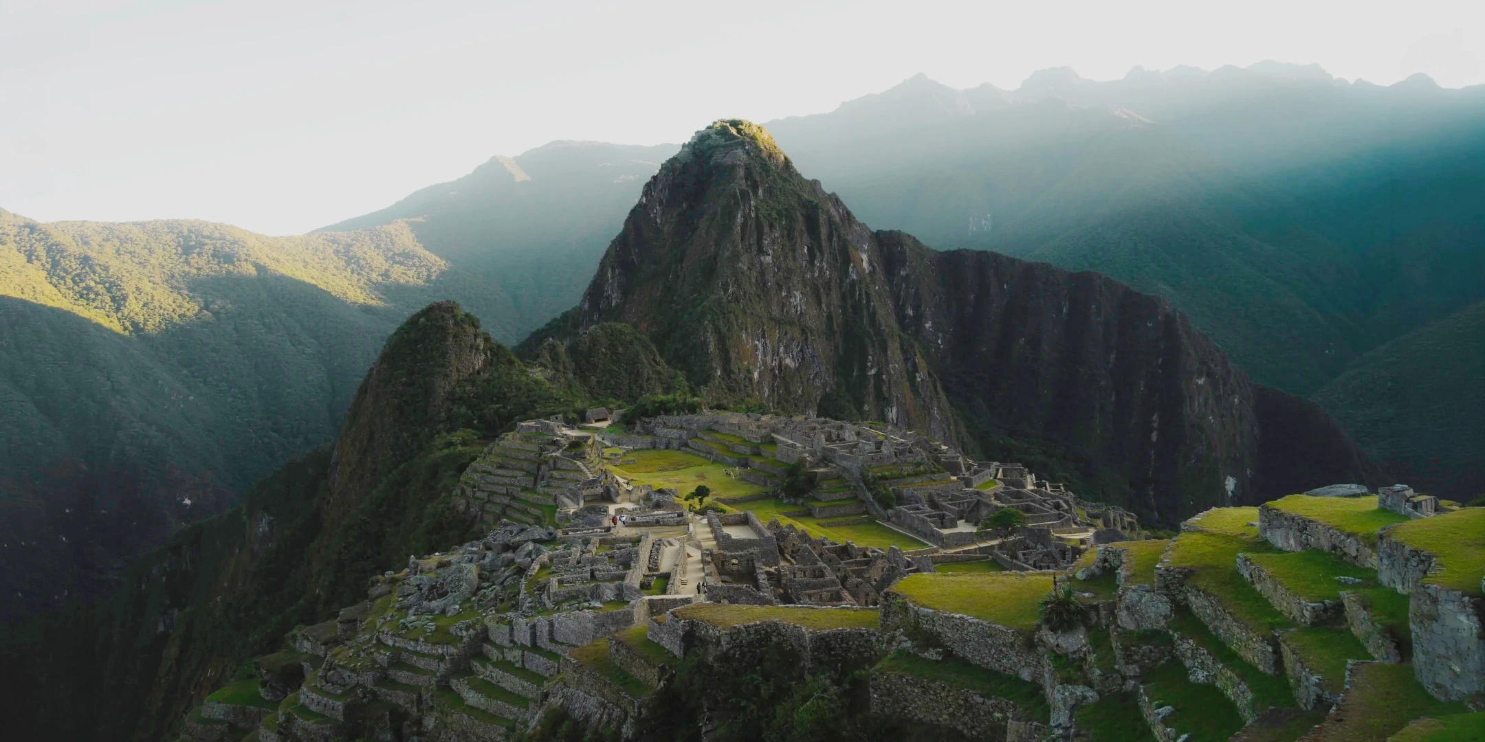High-angle panoramic view of the Machu Picchu sanctuary and Huayna Picchu mountain at sunrise.