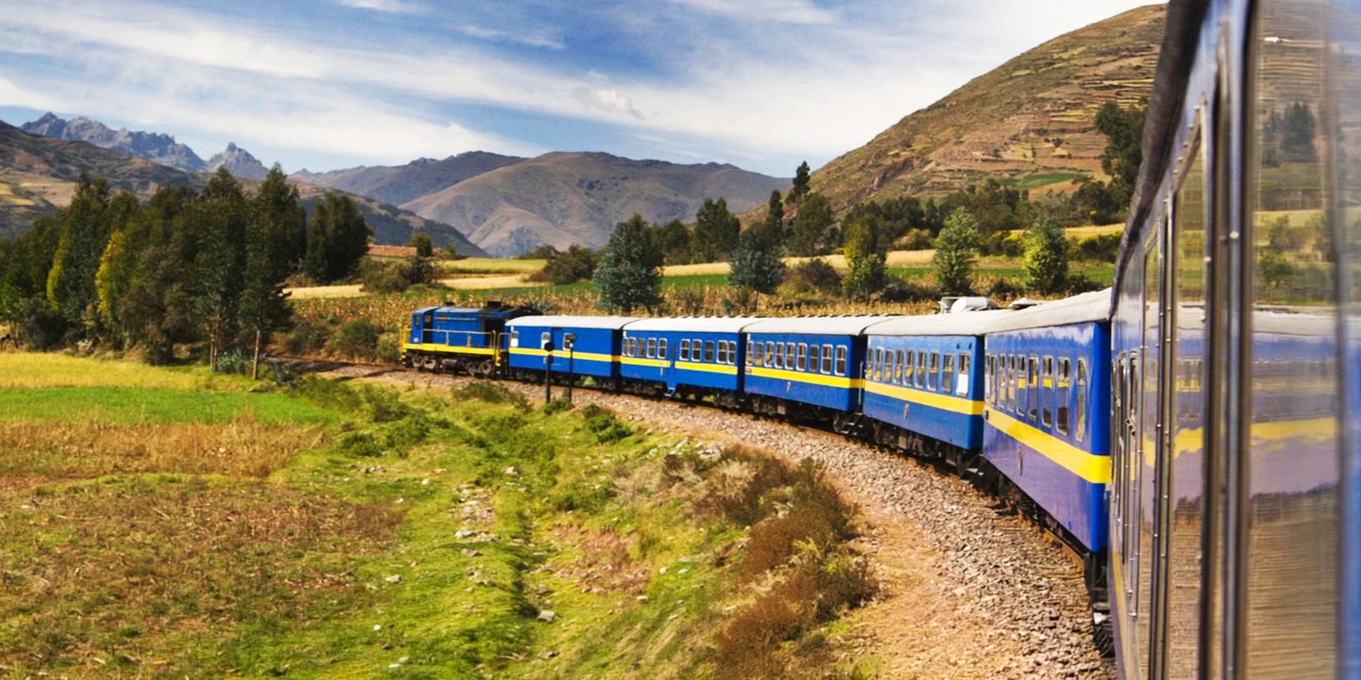 Wide shot of a blue and yellow train curving through the rural landscapes and agricultural fields of Cusco.
