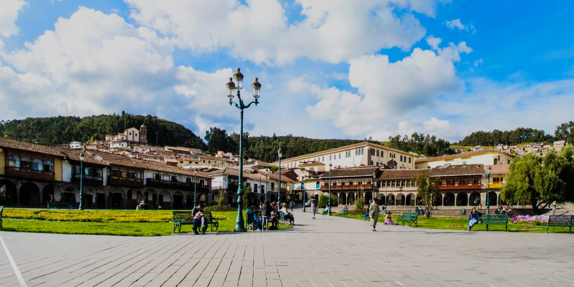 A wide view of Plaza Regocijo in Cusco with its central lamppost, surrounding colonial arches, and a view of the nearby hills under a cloudy sky.