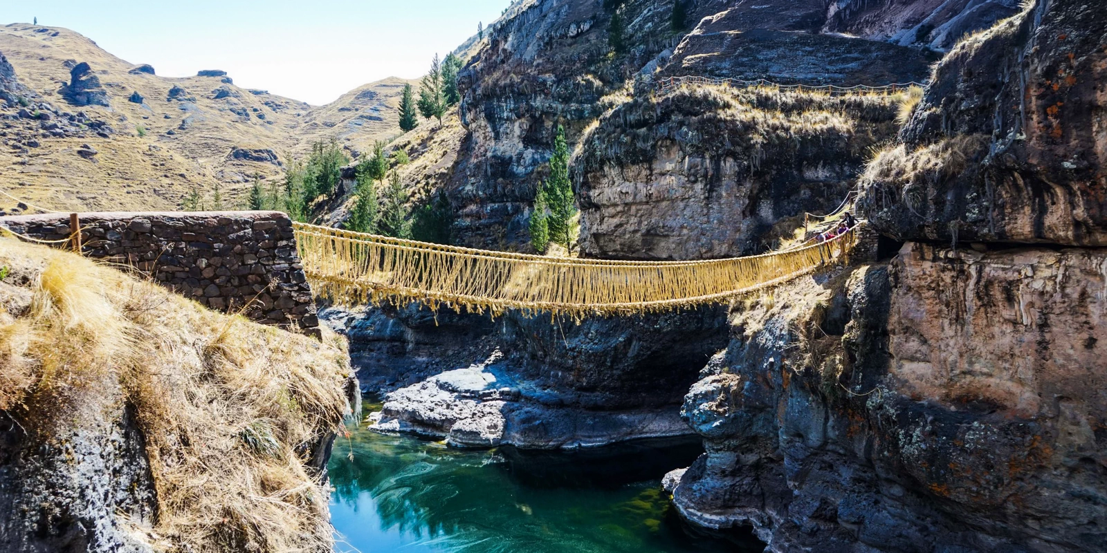 Golden grass rope bridge suspended over a turquoise river in the Peruvian Andes
