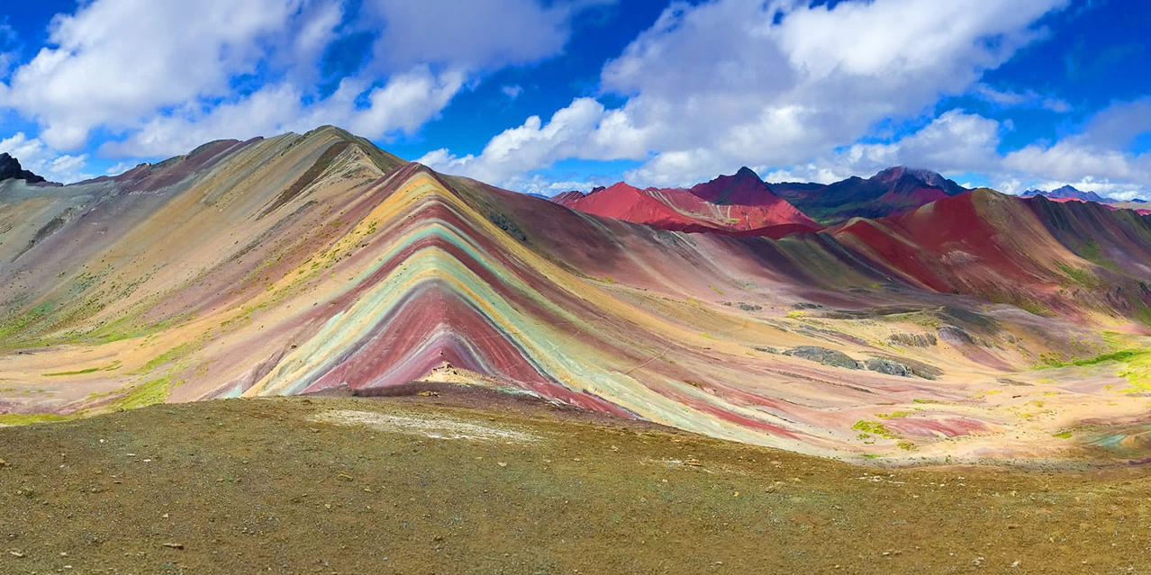 A wide panoramic view of the vibrant Rainbow Mountain in Peru with colorful mineral layers under a blue sky with white clouds.