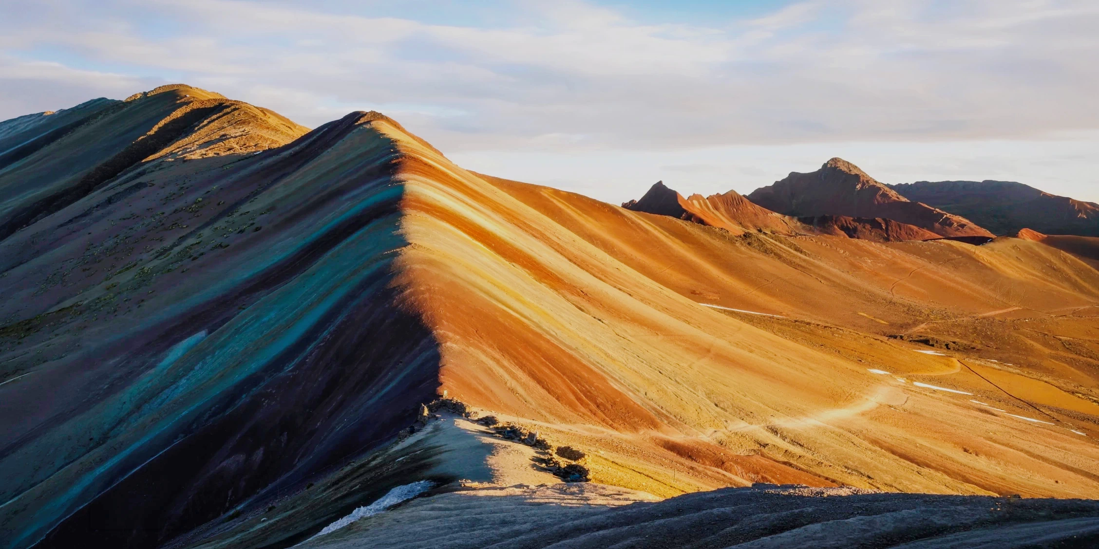 The colorful mineral layers of Rainbow Mountain under a soft morning light with trekking paths visible.
