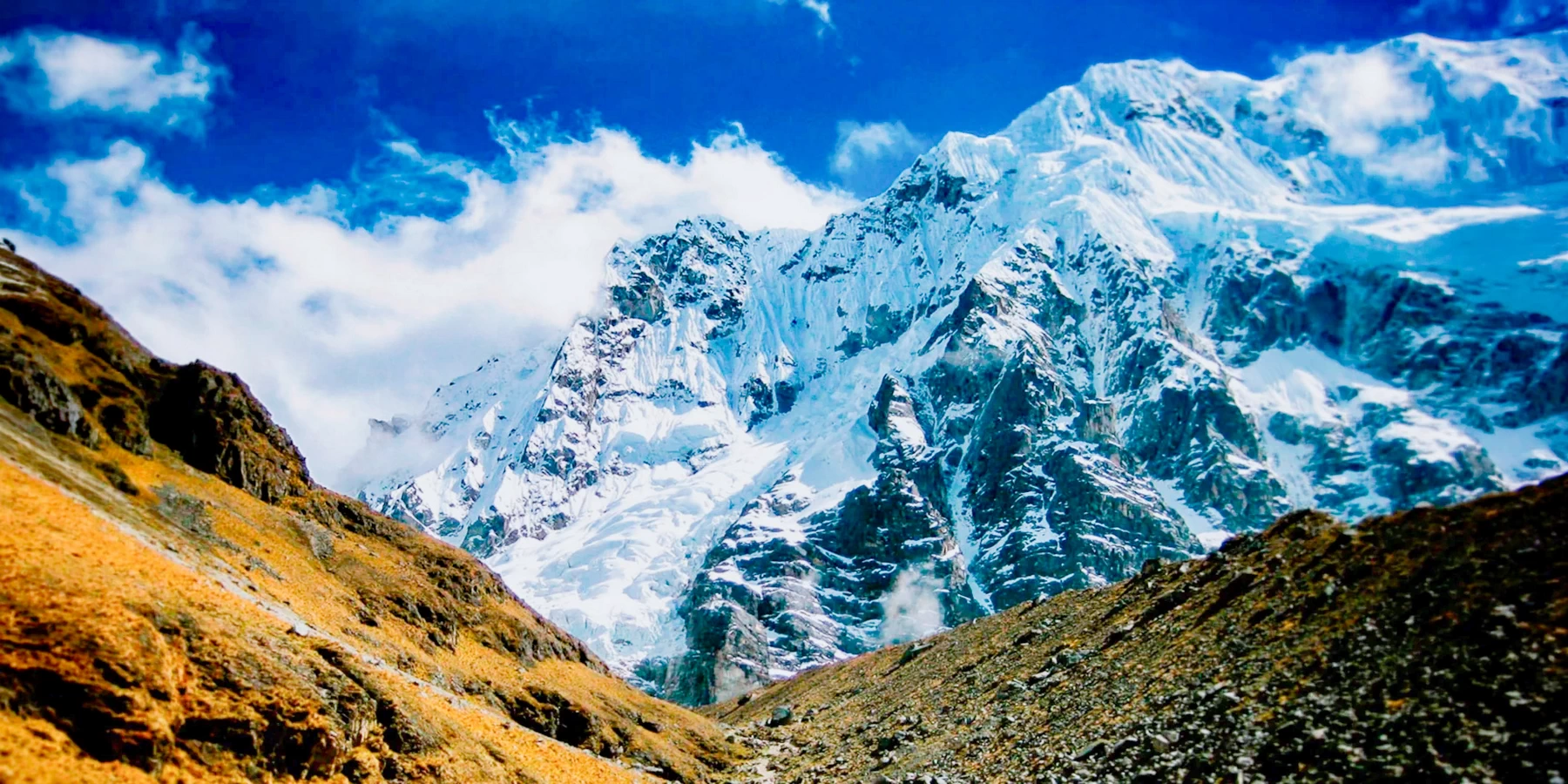 Panoramic view of the massive snow-covered Salkantay mountain under a bright blue sky with white clouds.
