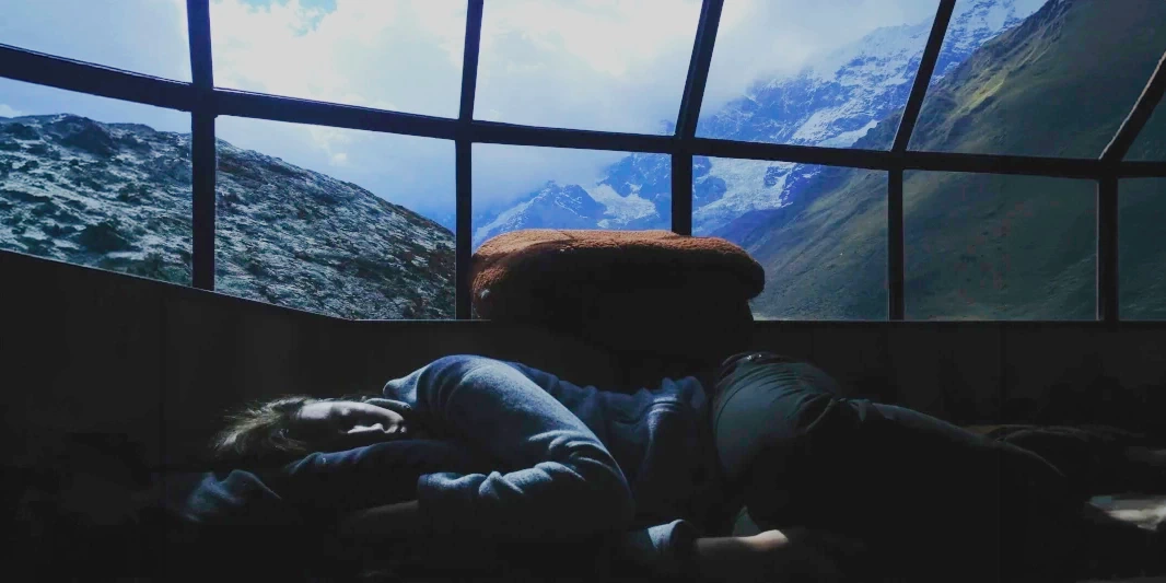 A person resting inside a glass-roofed dome with a panoramic view of the Andes mountains at dawn.