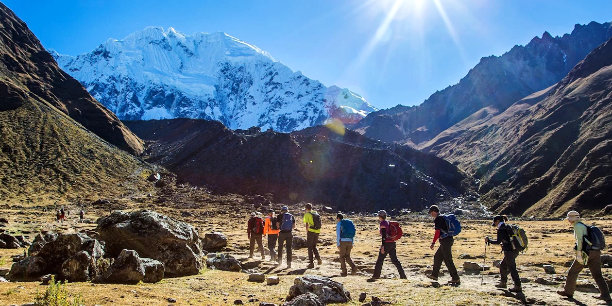 A line of hikers trekking through a wide mountain valley toward a massive white glacier under a bright sun.