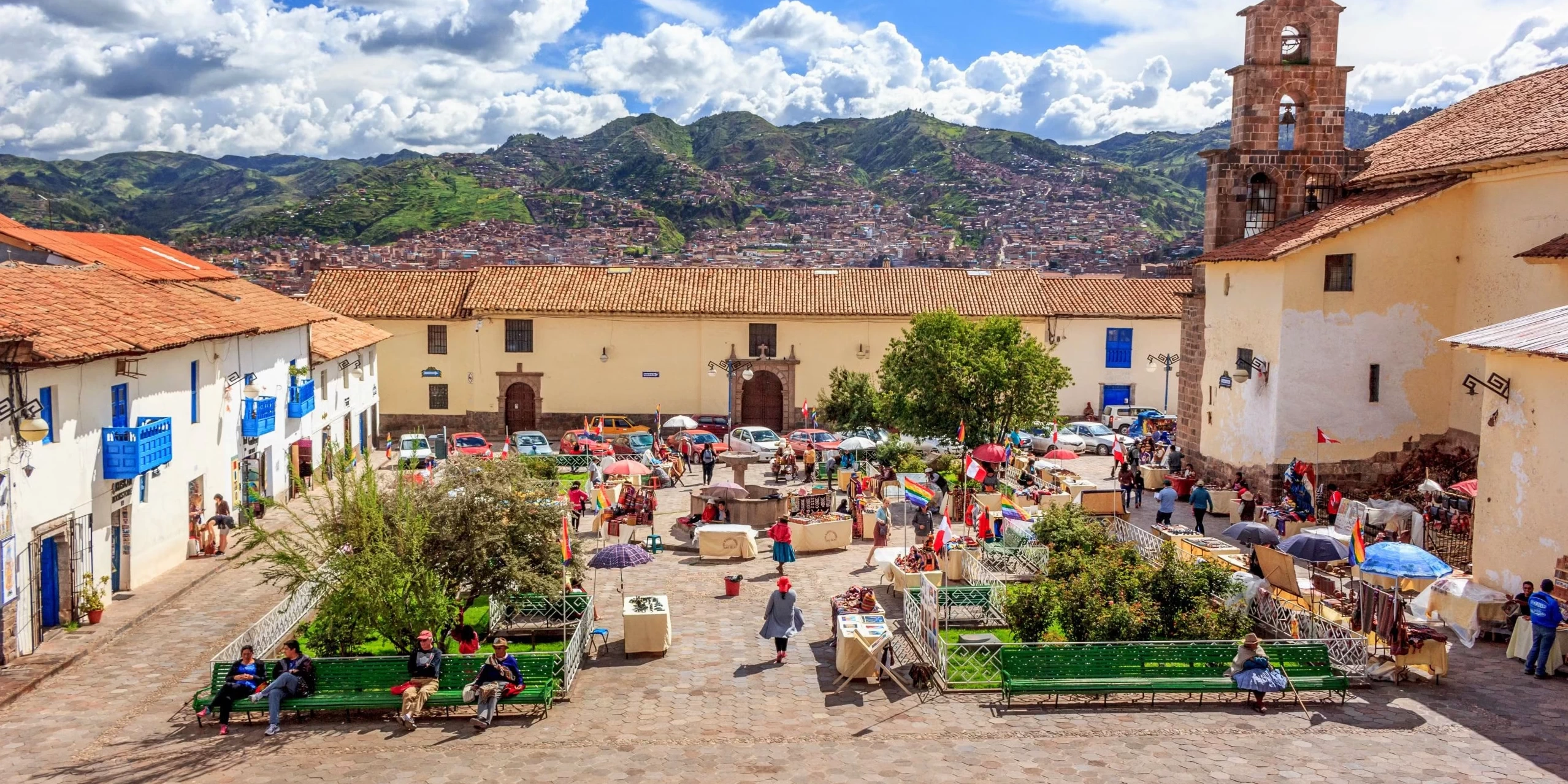 Panoramic view of San Blas square in Cusco featuring the colonial church, traditional white buildings with blue balconies, and local artisan stalls under a blue sky.