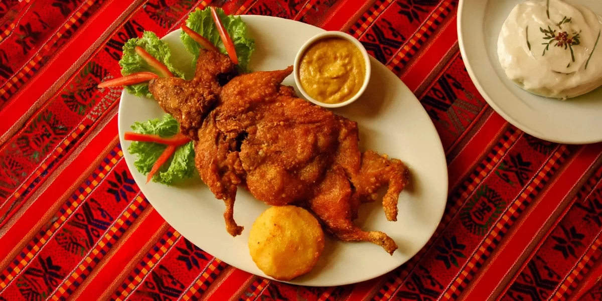 A crispy whole fried guinea pig known as Cuy Chactado, served with a golden potato and traditional Andean dipping sauce on a red patterned tablecloth.