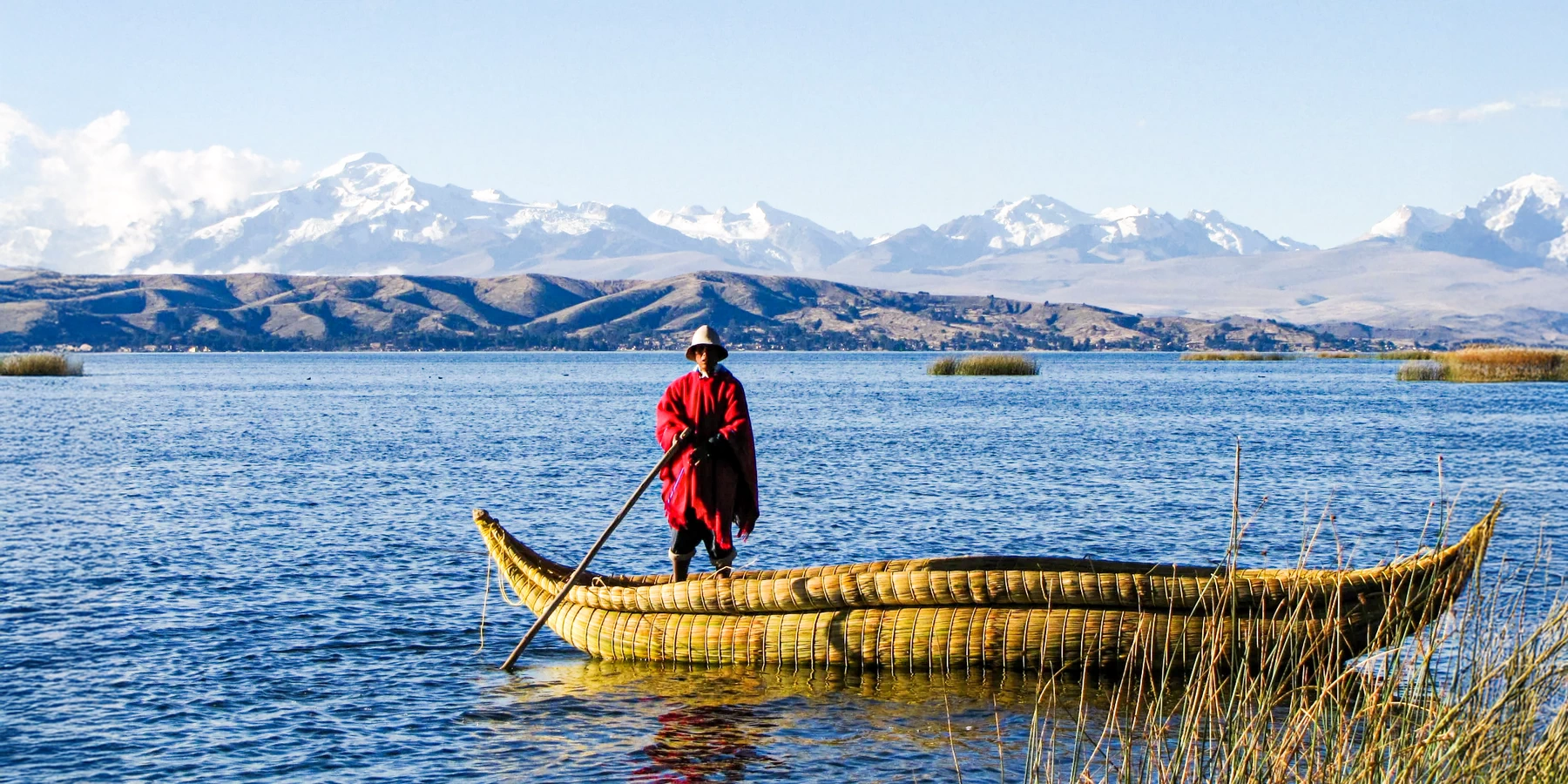 A local man in traditional red clothing rowing a totora reed boat on Lake Titicaca with snow-capped mountains in the back.
