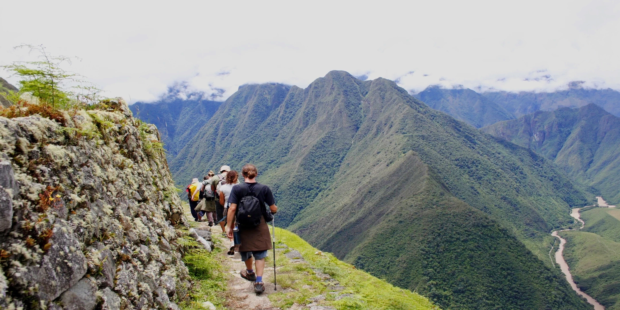 Tourists trekking along a narrow mountain ridge overlooking a deep green valley and a winding river in the Peruvian Andes.