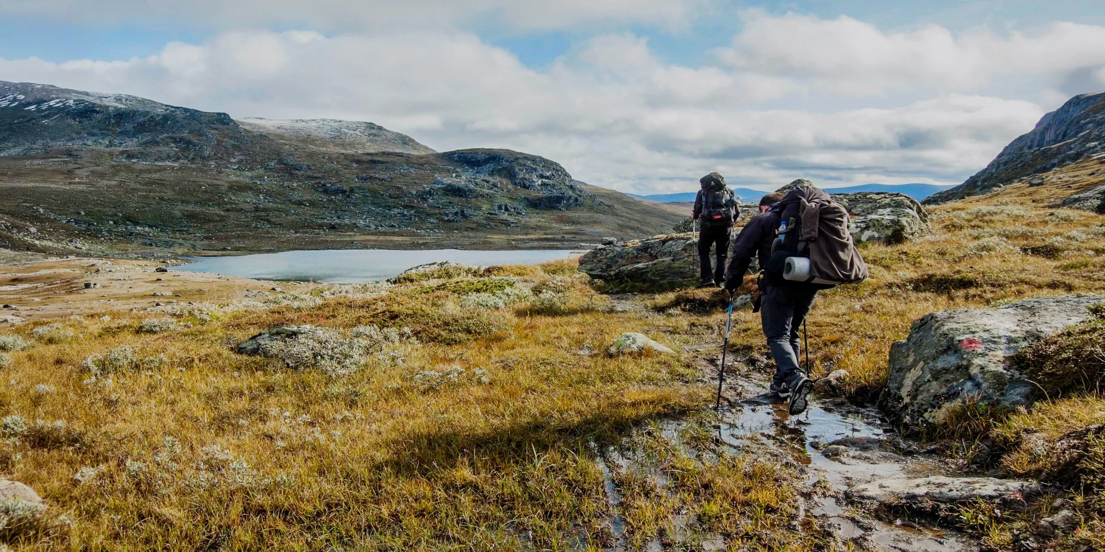 A hiker with a backpack standing on a rocky outcrop overlooking a deep blue mountain lagoon surrounded by green peaks.