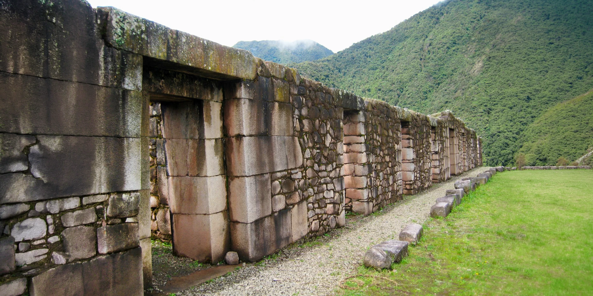 Long perspective of an ancient stone wall at the Vilcabamba archaeological site, surrounded by high mountains.