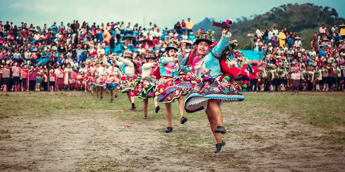 A group of dancers performing the Waynas dance in a large outdoor celebration with a crowd of spectators in the background.