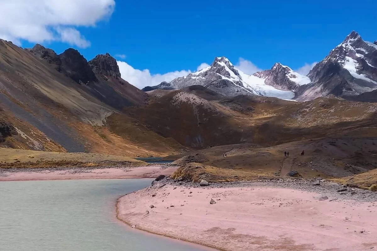 A breathtaking panoramic landscape of the Peruvian Andes during the Ausangate trek. It features a turquoise glacier lake, high altitude puna grasslands, and several snow-capped mountain ranges in the distance.