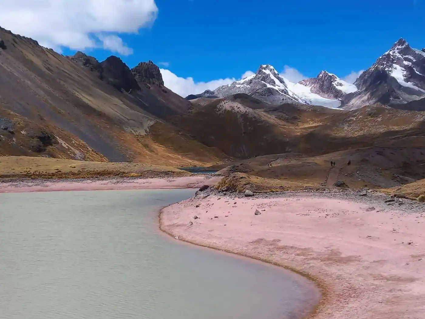 A breathtaking panoramic landscape of the Peruvian Andes during the Ausangate trek. It features a turquoise glacier lake, high altitude puna grasslands, and several snow-capped mountain ranges in the distance.
