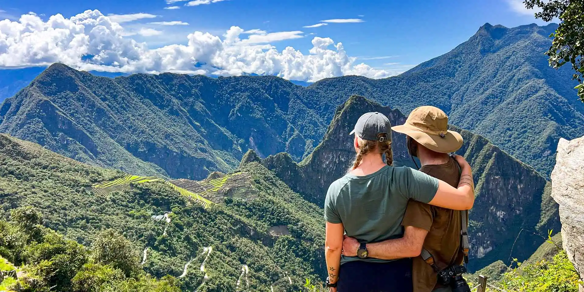 A young couple hugging while looking at the Machu Picchu Inca citadel and Huayna Picchu mountain from a viewpoint.