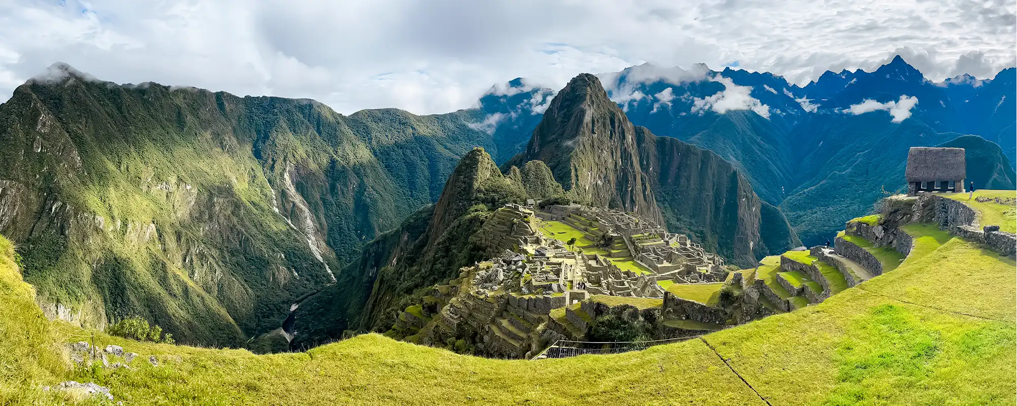 A wide panoramic landscape view of the Machu Picchu Inca citadel with the distinct Huayna Picchu mountain, green agricultural terraces, and dramatic Andean peaks under a partly cloudy sky.