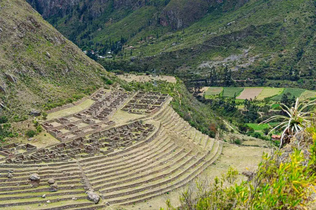 A stunning panoramic view of the original stone-paved Inca Trail winding its way up through a dramatic mountain pass. Dense Andean vegetation and other remote ruins are visible in the distance.
