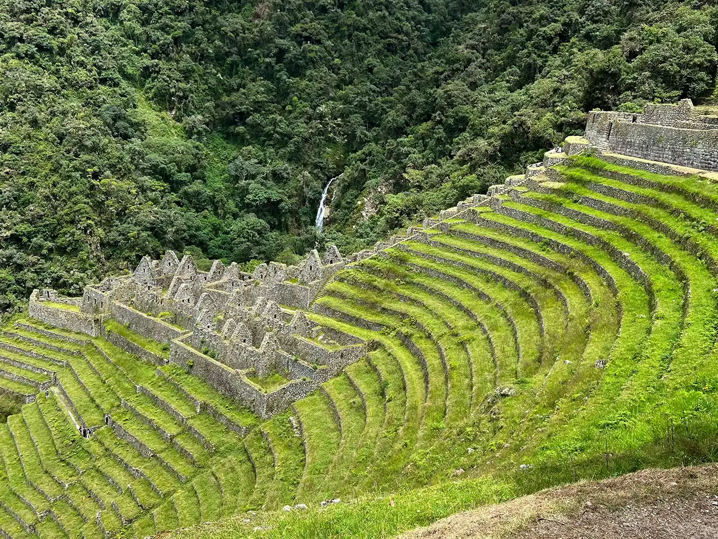 A dramatic high altitude Andean landscape with massive mountains and a winding river canyon. Part of the stone-paved Inca Trail is visible, leading towards Machu Picchu in the distance.