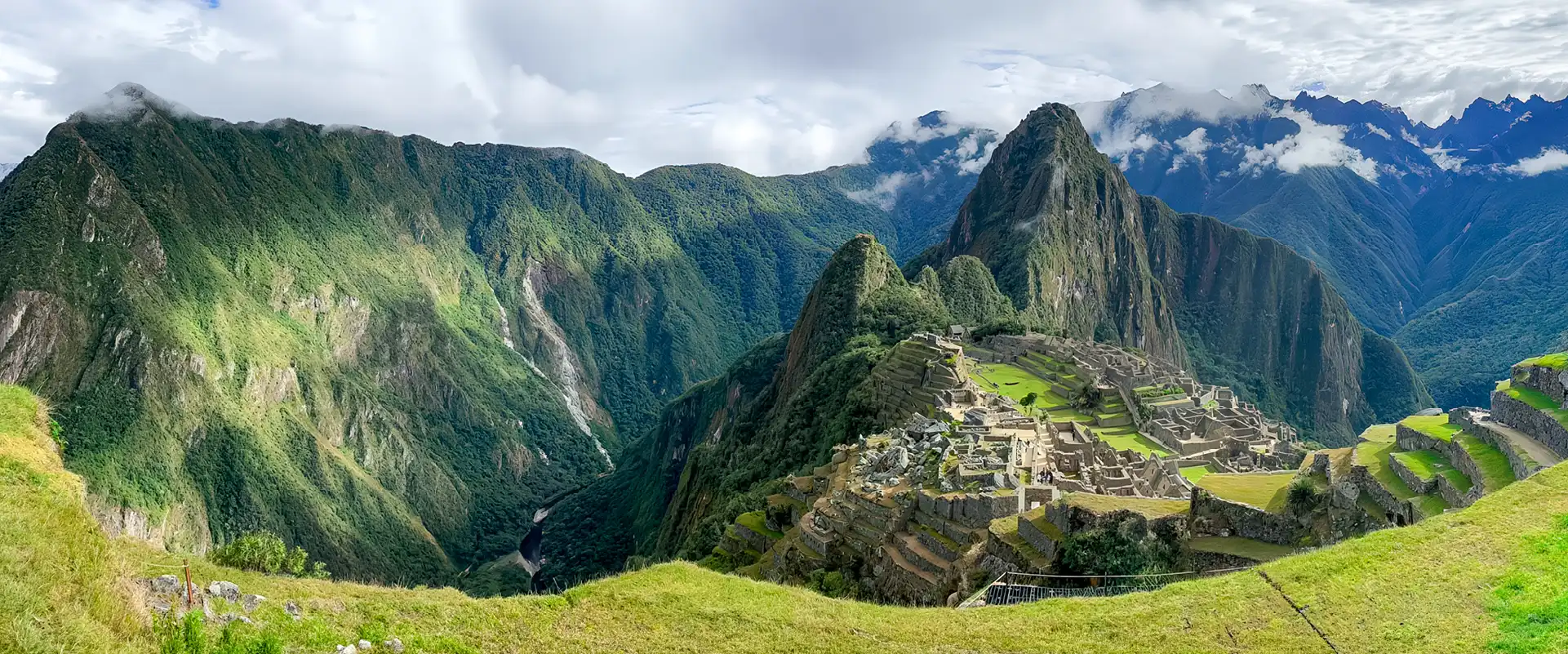 A wide panoramic landscape view of the Machu Picchu Inca citadel with the distinct Huayna Picchu mountain, green agricultural terraces, and dramatic Andean peaks under a partly cloudy sky.