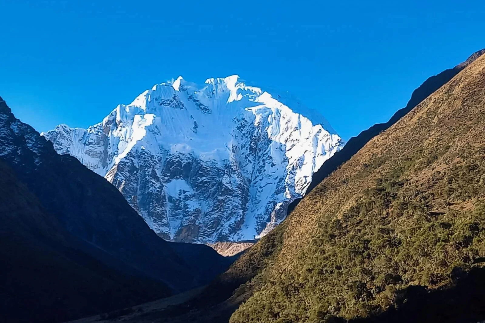 A sweeping panoramic landscape of the Salkantay trek. It features a massive snow-capped Salkantay nevado overlooking a dramatic high alpine valley with multiple glacier lakes and puna grasslands.
