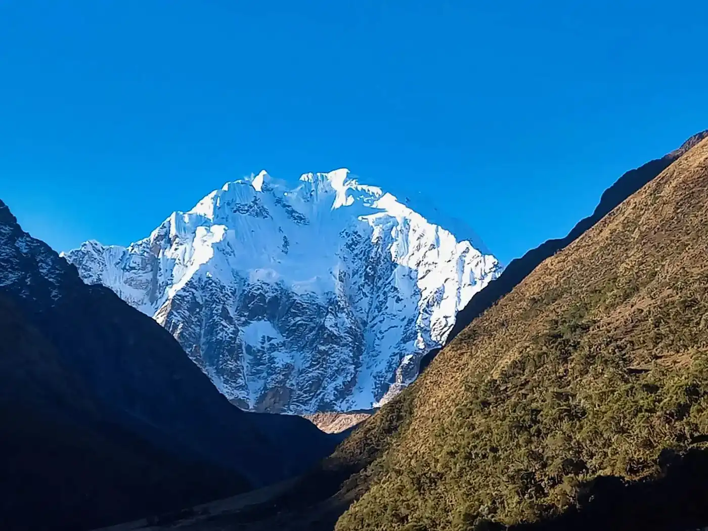 A sweeping panoramic landscape of the Salkantay trek. It features a massive snow-capped Salkantay nevado overlooking a dramatic high alpine valley with multiple glacier lakes and puna grasslands.