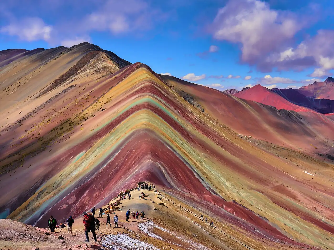 A vibrant panoramic view of the Vinicunca Rainbow Mountain in Peru. A single person is walking along the colorful ridge trail, emphasizing the scale of the majestic Andean peaks.