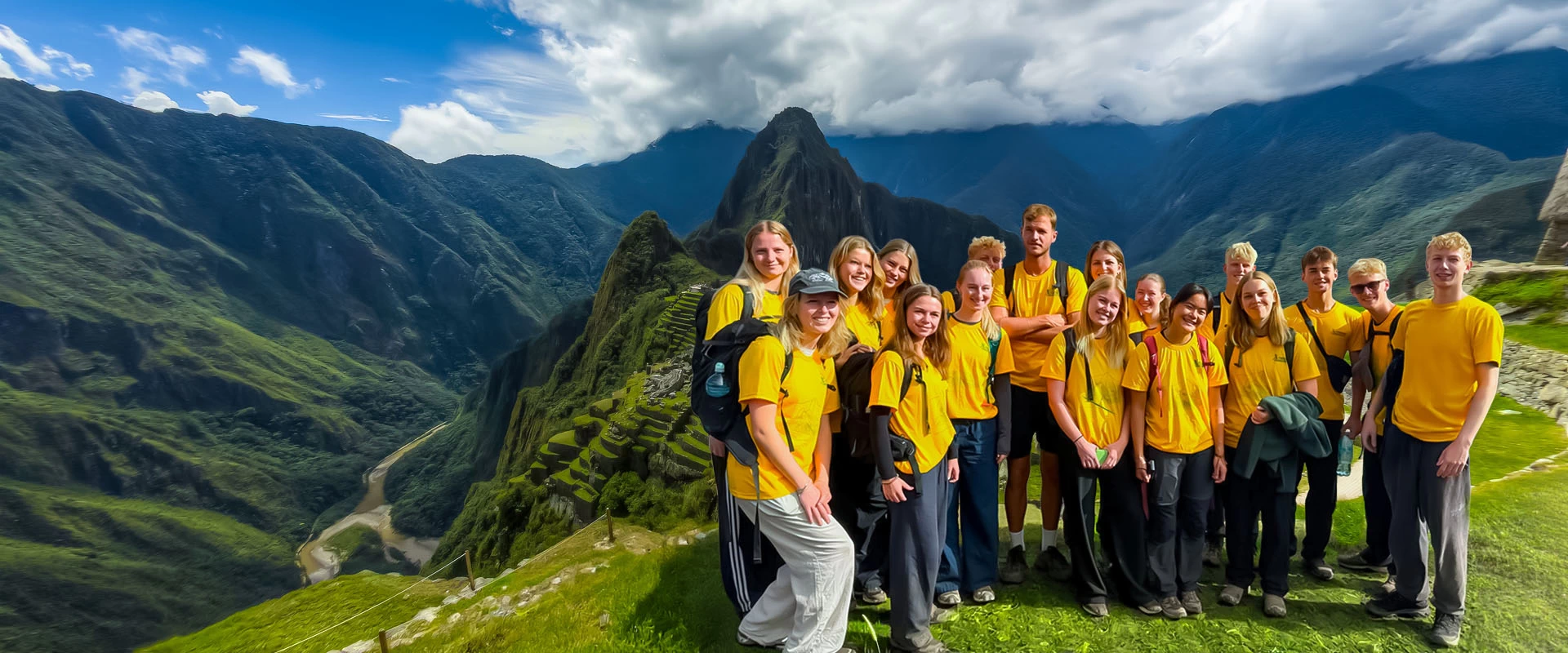 A large group of young travelers in yellow t-shirts smiling at the Machu Picchu citadel viewpoint with the Andes mountains in the background.