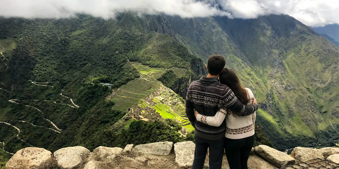 A couple wearing traditional alpaca sweaters stands embraced on a stone ledge, looking down at the panoramic view of the Machu Picchu citadel and the winding Hiram Bingham road from the summit of Huayna Picchu.