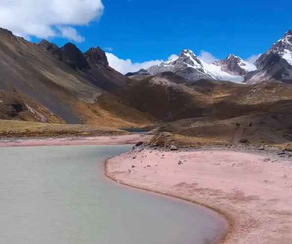 A breathtaking panoramic landscape of the Peruvian Andes during the Ausangate trek. It features a turquoise glacier lake, high altitude puna grasslands, and several snow-capped mountain ranges in the distance.