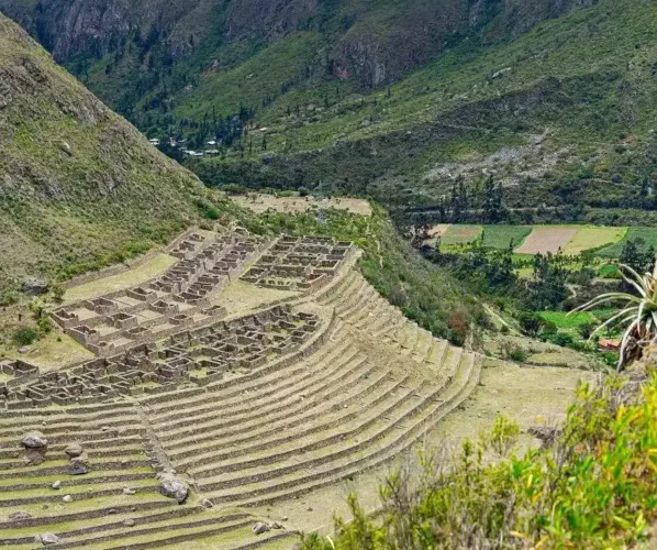 A stunning panoramic view of the original stone-paved Inca Trail winding its way up through a dramatic mountain pass. Dense Andean vegetation and other remote ruins are visible in the distance.