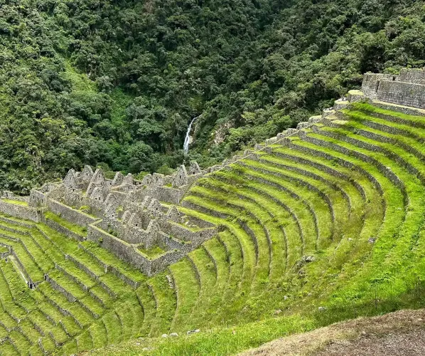 A dramatic high altitude Andean landscape with massive mountains and a winding river canyon. Part of the stone-paved Inca Trail is visible, leading towards Machu Picchu in the distance.
