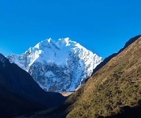A sweeping panoramic landscape of the Salkantay trek. It features a massive snow-capped Salkantay nevado overlooking a dramatic high alpine valley with multiple glacier lakes and puna grasslands.