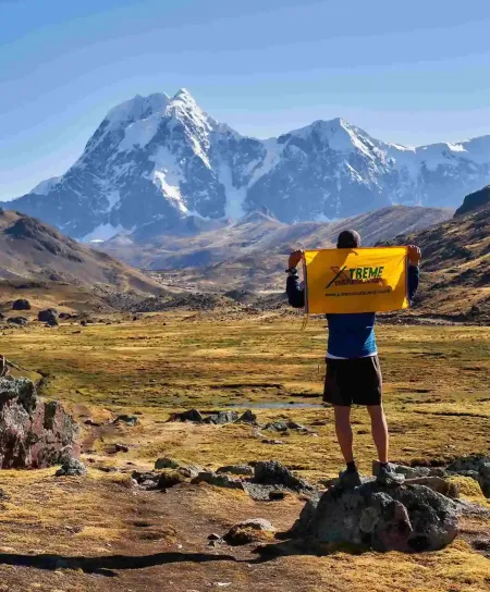 A majestic close-up shot of a prominent snow-capped Andean peak during the Ausangate trek in Peru. A lone traveler is walking on a winding trail below, highlighting the mountain's grand scale.