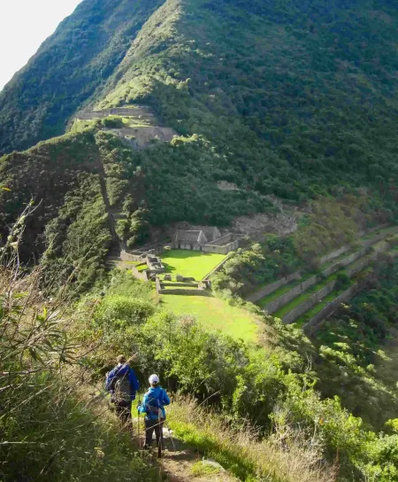 A magnificent long-distance panoramic overview of the remote Choquequirao archaeological park. Extensive stone terraces cling to a steep Andean mountain ridge overlooking a deep green valley and distant mountains under a cloudy sky. A small group of travelers is visible, highlighting its lack of crowds.